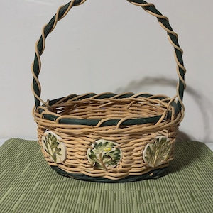 May include: Woven wicker basket with a green handle and trim. The basket features three decorative ceramic leaf designs around the base. The handle is wrapped with a twisted green and tan material. The basket sits on a green mat.