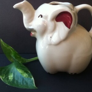 May include: A white ceramic pitcher shaped like an elephant with a trunk forming the spout and a handle on the back. The elephant features red accents inside the ears and a small red mouth. A green and yellow variegated leaf rests beside it.