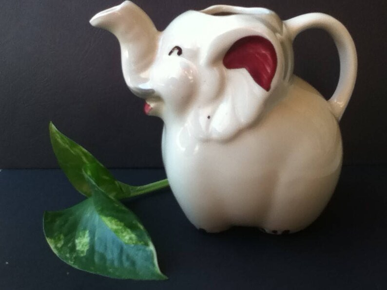 May include: A white ceramic pitcher shaped like an elephant with a trunk forming the spout and a handle on the back. The elephant features red accents inside the ears and a small red mouth. A green and yellow variegated leaf rests beside it.
