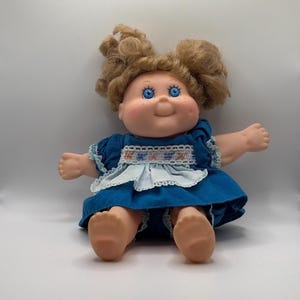 May include: A vintage doll with a round face, rosy cheeks, and blue eyes. The doll has light brown curly hair and is wearing a blue dress with white lace trim and a decorative apron. The doll is sitting with its arms outstretched.