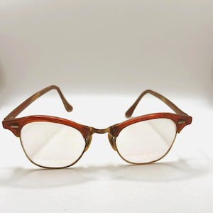 May include: Browline glasses featuring a reddish-brown tortoiseshell upper frame and thin gold-tone metal rims. The temples are a matching reddish-brown tortoiseshell pattern, and the bridge is a textured gold-tone metal. Clear lenses are included.