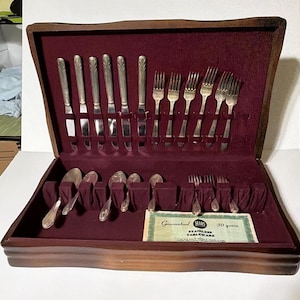 May include: A vintage silverware set in a wooden case with a burgundy velvet interior. The set includes knives, forks, and spoons, neatly arranged. A guarantee card from Sears is visible. The silverware has a decorative handle design.