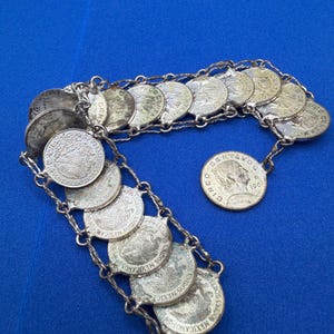 May include: A silver-toned bracelet made of Mexican coins. The coins are linked together with a delicate chain. The bracelet is displayed on a blue surface. The coins have text and images.