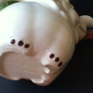May include: A cream-colored ceramic teapot shaped like an elephant. The teapot features a curved handle, a trunk-shaped spout, and small brown painted details on the feet. The surface is smooth and glossy, with a rounded body and a handle on the side.
