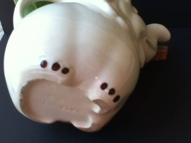 May include: A cream-colored ceramic teapot shaped like an elephant. The teapot features a curved handle, a trunk-shaped spout, and small brown painted details on the feet. The surface is smooth and glossy, with a rounded body and a handle on the side.