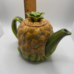 May include: A ceramic teapot shaped like a pineapple with a yellow and brown textured body, a green handle, and a green spout. The lid features a green pineapple crown. A wooden ruler is positioned behind the teapot, showing a height of 5 inches.