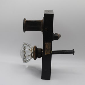 May include: Antique door hardware set featuring a clear, ribbed glass doorknob with a brass base. The set includes a black rectangular backplate, a bronze-colored handle, and a bolt. The hardware is designed for a door.