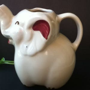 May include: A cream-colored ceramic pitcher shaped like an elephant. The pitcher features a trunk-shaped spout, a curved handle, and large ears with dark red accents on the inner portions. The elephant has small painted eyes and a mouth.