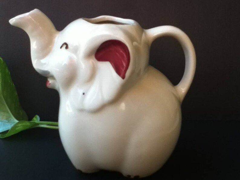 May include: A cream-colored ceramic pitcher shaped like an elephant. The pitcher features a trunk-shaped spout, a curved handle, and large ears with dark red accents on the inner portions. The elephant has small painted eyes and a mouth.