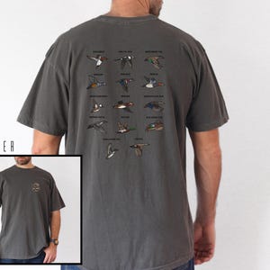 May include: Pepper-colored t-shirt featuring a detailed illustration of various duck species in flight. Each duck is labeled with its name. The shirt has a classic crew neck and short sleeves. The lower left corner shows a smaller image of the shirt.