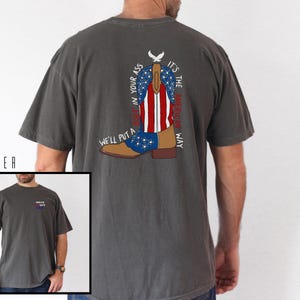 May include: Pepper-colored t-shirt with a graphic of a cowboy boot in red, white, and blue, with the text "It's the American Way" and "We'll put a boot in your ass". The shirt also has a small graphic on the front.