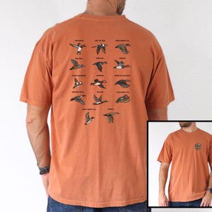 May include: Burnt orange t-shirt featuring illustrations of various duck species in flight, with their names above each image. The shirt also has a small graphic on the front. The brand logo "E & L DESIGNS" is visible on the bottom left.