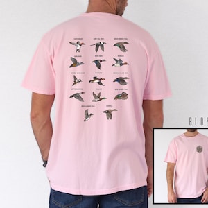 May include: A light pink t-shirt featuring a detailed illustration of various duck species in flight, with their names above them. The shirt also has a small emblem on the front. The text "BLOSSOM" is visible on the right side.