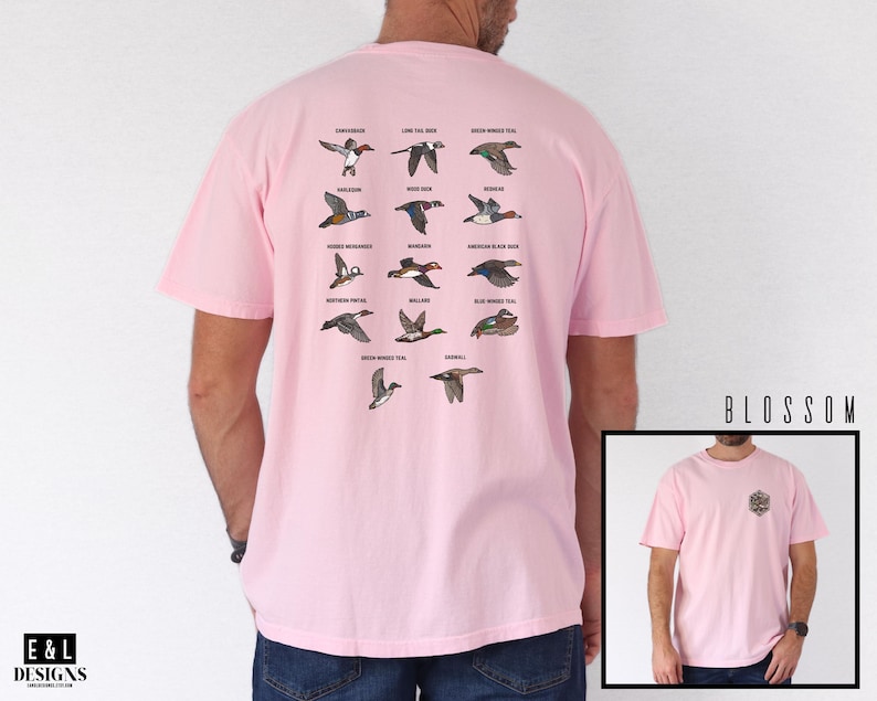May include: A light pink t-shirt featuring a detailed illustration of various duck species in flight, with their names above them. The shirt also has a small emblem on the front. The text "BLOSSOM" is visible on the right side.