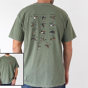 May include: Moss green t-shirt featuring a detailed illustration of various duck species in flight. The back of the shirt displays a grid of colorful duck illustrations with their names. The shirt is a comfortable, casual style.