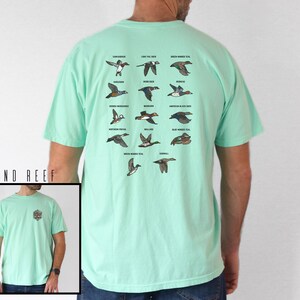 May include: A light green t-shirt featuring a detailed illustration of various duck species. The back of the shirt displays a grid of duck illustrations with their names. The shirt also has a small logo on the front. The brand name "E&L DESIGNS" is visible.