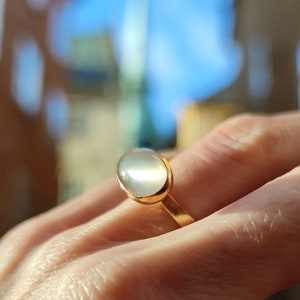 Könnte beinhalten: Ein goldener Ring mit einem weißen ovalen Edelstein. Der Ring befindet sich an einem Finger.