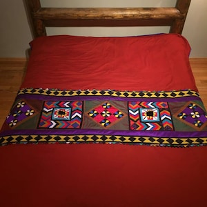 May include: A red bedspread with a colorful patterned band across the bottom. The band features geometric shapes and designs in shades of red, yellow, blue, green, and purple.