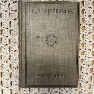 May include: A vintage copy of "Les Misérables" by Victor Hugo. The book has a muted brown cover with the title and author's name in dark lettering. A decorative emblem is centered on the cover. The book is resting on a cream-colored crocheted surface.