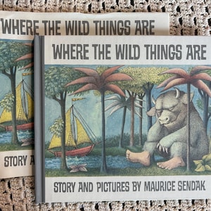 May include: Two copies of the hardcover children's book Where the Wild Things Are by Maurice Sendak. The cover features a grey monster sitting under a palm tree near a red and yellow sailboat on water, with the title printed in bold black letters.