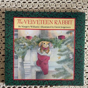 May include: A hardcover book titled The Velveteen Rabbit by Margery Williams, illustrated by David Jorgensen. The cover features a brown rabbit inside a red stocking hanging on a fireplace mantel with greenery, candles, and a Christmas tree.