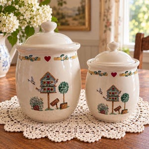 May include: Two cream-colored ceramic canisters with matching lids, featuring a painted garden scene with a blue birdhouse, green topiary, small red hearts, and flying birds. The canisters have a rounded shape and a glossy finish.