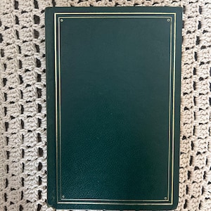 May include: A dark green photo album with a gold border. The album is rectangular and appears to be closed. The background is a cream-colored, crocheted surface.