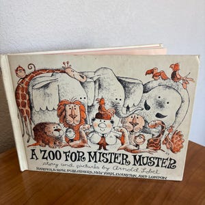 May include: A hardcover children's book titled A ZOO FOR MISTER MUSTER, story and pictures by Arnold Lobel, published by Harper & Row. The cover features a black and white line drawing with orange accents depicting a man surrounded by zoo animals.