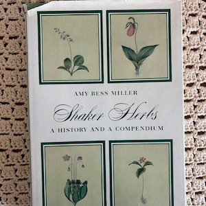 May include: A hardcover book titled Shaker Herbs: A History and a Compendium by Amy Bess Miller. The white cover features four rectangular illustrations of botanical plants in green frames, with black text centered on the front.