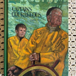 May include: A hardcover book titled Captains Courageous by Rudyard Kipling. The cover features an illustration of an older person and a younger person wearing yellow rain jackets, standing at a ship's wooden steering wheel against a green sea.
