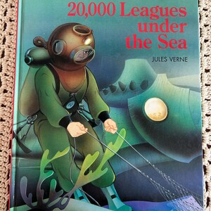 May include: A vintage hardcover book titled "20,000 Leagues Under the Sea" by Jules Verne. The cover features an illustration of a diver in a copper helmet and green suit, with a submarine in the background. The book is set against a beige crocheted background.