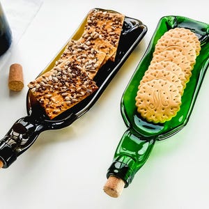 Pode incluir: Dois pratos de servir de garrafa de vidro, um verde e outro castanho, com uma borda decorativa. O prato verde tem uma pilha de biscoitos redondos, e o prato castanho tem uma pilha de bolachas retangulares com sementes.