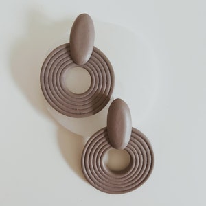 May include: A pair of taupe-colored earrings. Each earring features a rounded oval top connected to a circular pendant with concentric grooves. The earrings are displayed on a white surface.