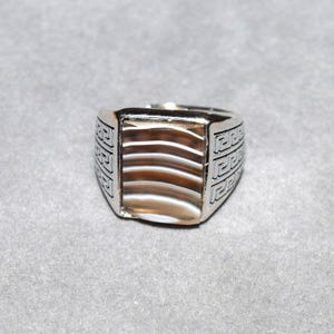 May include: A silver-toned ring featuring a rectangular center stone with horizontal brown and white banded layers. The metal band is decorated with a repeating geometric Greek key pattern on both sides of the central stone setting.