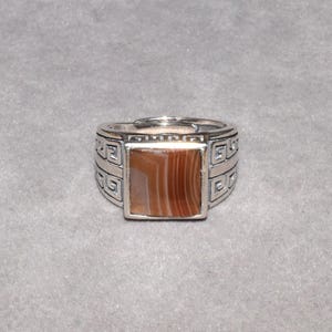 May include: A silver-toned ring featuring a square-shaped, brown and white banded agate stone inlay. The metal band is decorated with a repeating geometric Greek key pattern on both sides and includes an adjustable opening at the back.