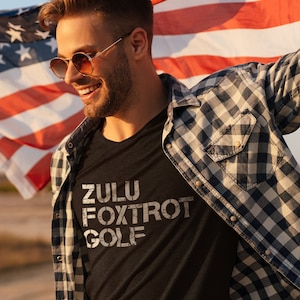 May include: A man wearing a black t-shirt with the words "ZULU FOXTROT GOLF" printed in white. He is also wearing a blue and white plaid shirt and sunglasses. He is holding an American flag.