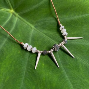 May include: A silver necklace with white and grey beads and silver spikes. The necklace is on a green leaf.