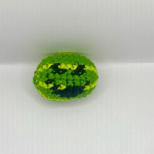 May include: A handmade, crocheted watermelon-shaped toy. The toy features a green exterior with yellow stripes and a dark green pattern resembling seeds. The item is likely a pet toy or a decorative item.