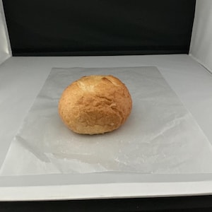May include: A golden-brown, round loaf of bread, possibly a roll or bun, sits on a sheet of white parchment paper. The bread has a textured crust and appears freshly baked. The image is taken in a studio setting with a white background.