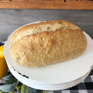 May include: A loaf of freshly baked bread rests on a white, distressed cake stand. The bread has a golden-brown crust and a textured surface. The cake stand is placed on a black and white checkered tablecloth. A wooden sign with the words "pies, cakes, pastries" is in the background.