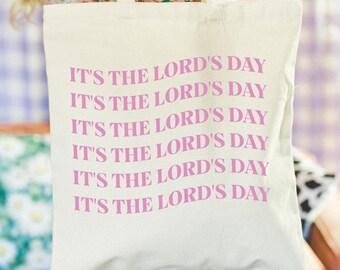 Può includere: Una borsa tote in tela bianca con la frase "It's the Lord's Day" ripetuta sei volte in lettere rosa.