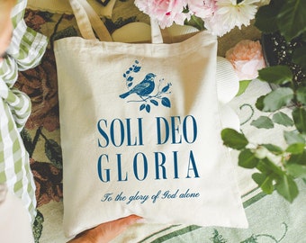Può includere: Una borsa tote color crema con un design di uccello blu e floreale, con le parole "SOLI DEO GLORIA" e "Alla gloria di Dio solo" stampate sul davanti. La borsa ha lunghe cinghie ed è tenuta da una persona.