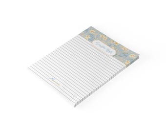 May include: A rectangular notepad with horizontal black lines on a white background. The top section features a blue floral pattern with yellow flowers and a central white label containing the text Scripture Notes in blue script. A small logo is visible.