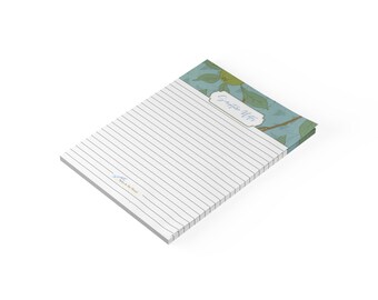 May include: A rectangular notepad with horizontal lines for writing. The top section features a blue background with green leaf illustrations and a white label containing the text Scripture Notes. A small logo is printed in the bottom left corner.