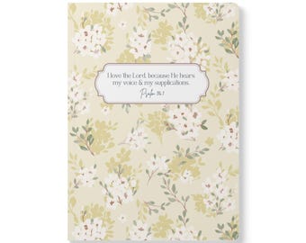 May include: A rectangular notebook with a light beige background featuring a repeating pattern of white flowers and green leaves. Centered on the cover is a white label with a thin border containing the text: I love the Lord, because He hears my voice & my supplications. Psalm 116:1.