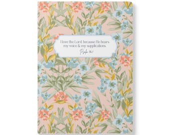 May include: A rectangular notebook with a light pink background featuring a repeating floral pattern in shades of coral, light blue, and olive green. A central white label contains the text: I love the Lord, because He hears my voice & my supplications. Psalm 116:1.