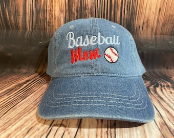 May include: A denim baseball cap with the words "Baseball Mom" embroidered in white and red. A baseball graphic is on the right side. The hat has a curved brim and adjustable strap.