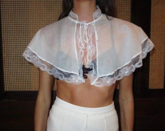 May include: A sheer white capelet featuring a high collar with a tie closure at the neck. The garment is constructed from a lightweight, semi-transparent fabric and is finished with a decorative scalloped lace trim along the entire bottom edge.