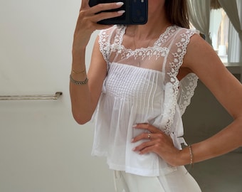 May include: A white sleeveless blouse featuring a square neckline, a pleated bodice, and sheer mesh shoulder panels adorned with intricate white floral embroidery and scalloped edges. The garment has a relaxed fit and a straight hemline.