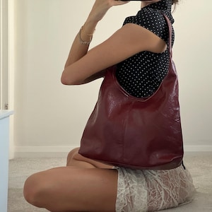 May include: A burgundy leather shoulder bag with a curved design. The bag is held by a person wearing a black top with white polka dots. The person is sitting on a light-coloured carpet.